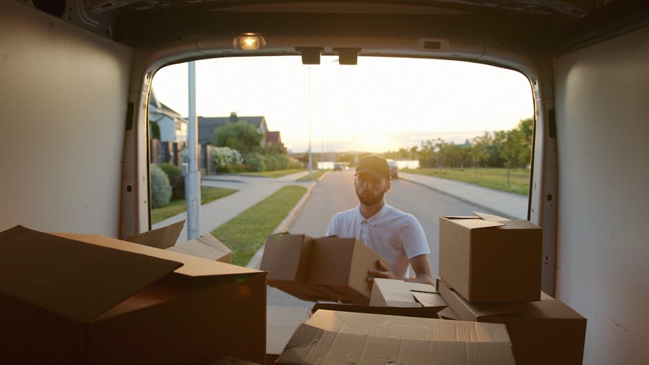 A man wearing a white polo shirt, cap, and glasses is seen through the open back of a moving van, loading it with various cardboard boxes of different sizes, some sealed with packing tape and others partially open. The boxes are placed inside the van, which is parked on a residential street with a sidewalk, grassy areas, and houses visible in the background. The photograph is taken during sunset, casting warm light on the scene and creating a serene atmosphere. The man appears to be engaged in a home relocation task, carefully handling the boxes as part of furniture transport and packing and moving process, with the van's interior showing signs of previous use for removals. This scene exemplifies the loading process involved with house removals, with the focus on efficient organization and safe handling of packing materials as part of professional removal services by Man With a Van Coulsdon.