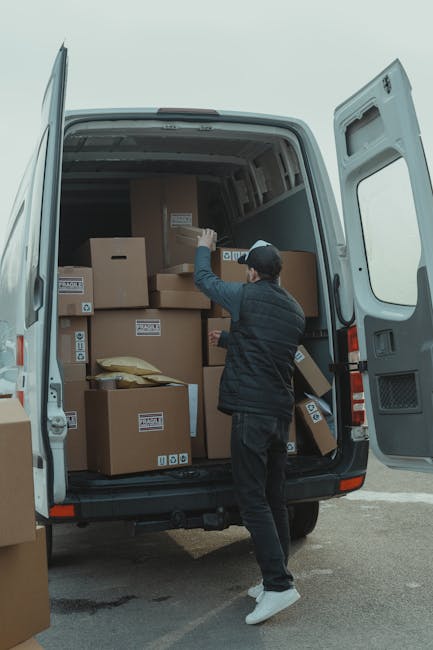 A man wearing a black jacket, dark pants, white sneakers, and a cap is inside the rear of a loaded white van with open sliding doors, arranging and handling cardboard boxes used for a home relocation. The boxes vary in size and are sealed with packing labels, some wrapped in plastic or placed on padded packing materials. The van is parked outside on a paved surface, with the pavement visible beneath the vehicle. The interior of the van is lined with dark panels, and the loading process appears organized, with boxes stacked systematically to optimize space. Nearby, folding trolleys and straps might be partially visible, indicating the use of equipment to facilitate furniture transport and packing during this moving process. This scene reflects the logistics involved in house removals and professional moving services, with a focus on careful handling and efficient loading by Man With a Van Coulsdon.