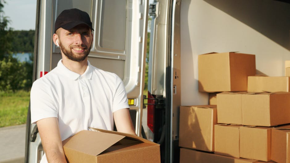 A man wearing a white polo shirt, cap, and glasses is seen through the open back of a moving van, loading it with various cardboard boxes of different sizes, some sealed with packing tape and others partially open. The boxes are placed inside the van, which is parked on a residential street with a sidewalk, grassy areas, and houses visible in the background. The photograph is taken during sunset, casting warm light on the scene and creating a serene atmosphere. The man appears to be engaged in a home relocation task, carefully handling the boxes as part of furniture transport and packing and moving process, with the van's interior showing signs of previous use for removals. This scene exemplifies the loading process involved with house removals, with the focus on efficient organization and safe handling of packing materials as part of professional removal services by Man With a Van Coulsdon.