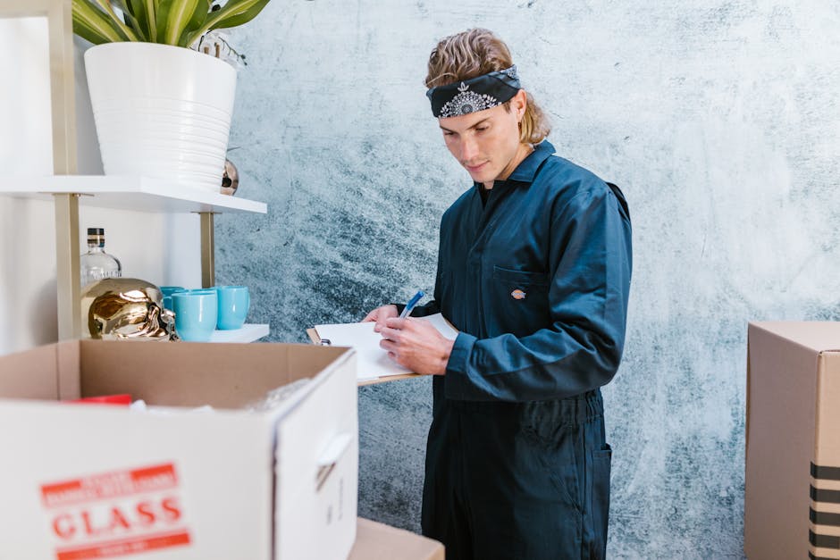 A young man wearing a dark blue jumpsuit and a black bandana with white patterns is inside a room with a textured light blue wall. He is holding a clipboard and pen, appearing to record information about the packing or moving process. In front of him, there are cardboard boxes, one with a red label, containing household items or packing materials. To his left, a white shelving unit displays decorative blue cups, a gold-colored object, and a clear glass bottle. On the top shelf, there is a large white ceramic pot with a green plant. The scene suggests a home relocation or packing activity as part of a furniture transport or moving service, with natural lighting and a tidy environment, consistent with the procedures of Man With a Van Coulsdon for house removals and safety compliance during loading and transport.