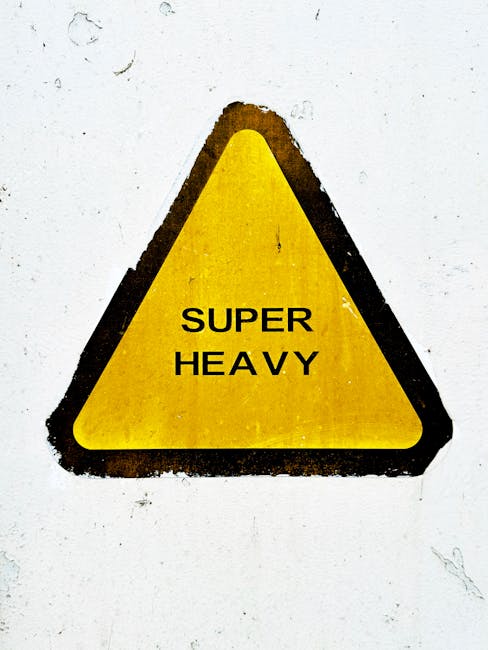 A yellow triangular warning sign with black border and bold black text reading 'SUPER HEAVY' is affixed to a white wall, indicating the presence of heavy items requiring careful handling. The sign is slightly worn with minor dirt marks around its edges. In the context of home relocation and furniture transport, this sign would typically be used to alert movers to the need for appropriate lifting equipment and safety procedures. The environment appears to be an interior or exterior area adjacent to a property, possibly near a doorway or loading zone where heavy furniture or appliances are being moved. This visual emphasizes the importance of safe moving practices, such as employing proper lifting techniques or using trolleys and straps, as part of professional removals services, like those provided by Man With a Van Coulsdon, ensuring efficient and secure packing and loading during the moving process.