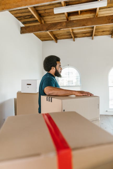 A young man wearing a dark blue jumpsuit and a black bandana with white patterns is inside a room with a textured light blue wall. He is holding a clipboard and pen, appearing to record information about the packing or moving process. In front of him, there are cardboard boxes, one with a red label, containing household items or packing materials. To his left, a white shelving unit displays decorative blue cups, a gold-colored object, and a clear glass bottle. On the top shelf, there is a large white ceramic pot with a green plant. The scene suggests a home relocation or packing activity as part of a furniture transport or moving service, with natural lighting and a tidy environment, consistent with the procedures of Man With a Van Coulsdon for house removals and safety compliance during loading and transport.
