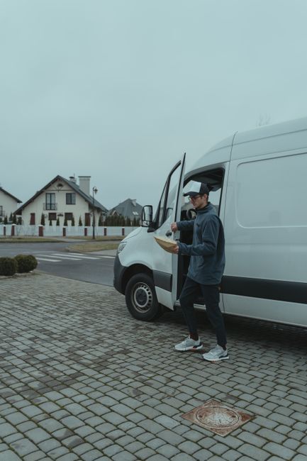 A male worker from Man With a Van Coulsdon stands beside a white cargo van parked on a paved driveway outside a residential property, preparing for a home relocation or furniture transport. He is dres