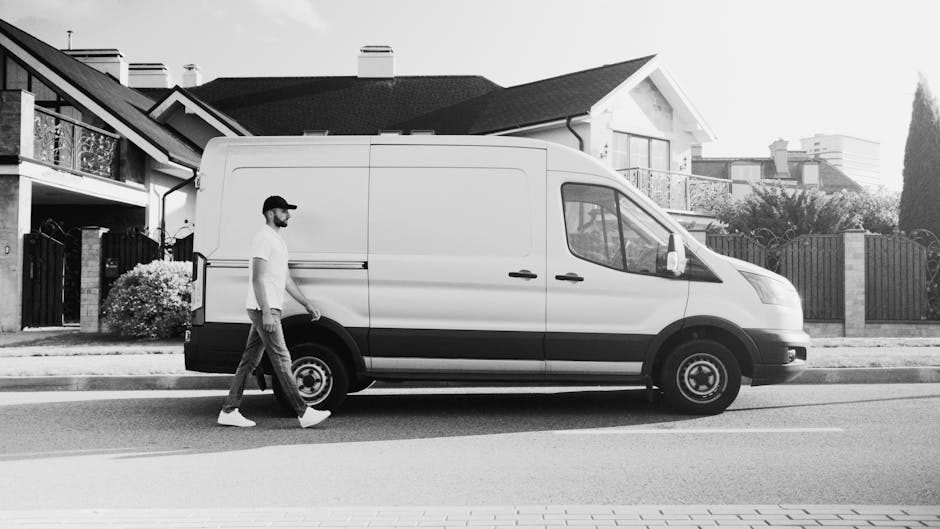 A male worker from Man With a Van Coulsdon stands beside a white cargo van parked on a paved driveway outside a residential property, preparing for a home relocation or furniture transport. He is dres