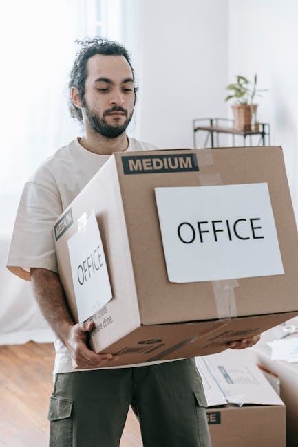 A man with dark, curly hair and a beard, dressed in casual light-colored clothing, is inside a bright, sparsely furnished room with white walls and natural light. He is holding a large cardboard box labeled 'OFFICE' with a smaller label indicating 'MEDIUM' size, as part of a home or office relocation process. In the background, there is a small table with a potted plant on top, and some additional packaging materials and boxes are visible on the floor. The image illustrates the packing and loading process during a house or office move, with the man representing professional removals services such as those offered by Man With a Van Coulsdon, specializing in furniture transport and packing for local relocations.