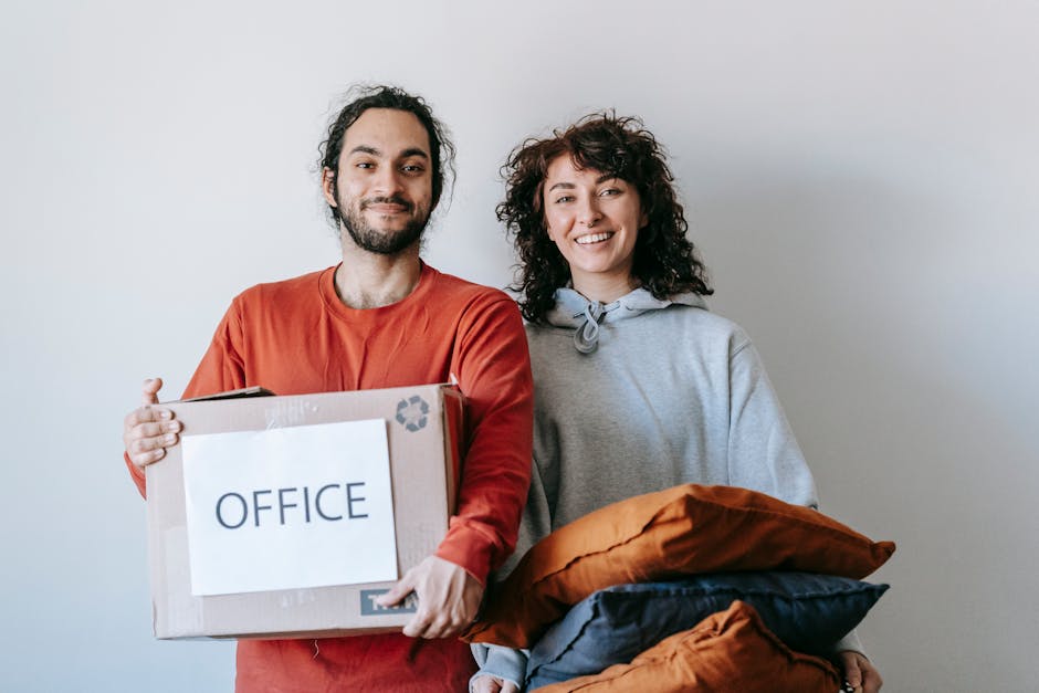 A man and a woman standing indoors against a plain white wall, preparing for a house relocation. The man, with dark curly hair and a beard, is holding a cardboard box labeled 'OFFICE' with a recycling symbol, indicating packing for moving. He is dressed in a red-orange long-sleeve shirt. The woman, with shoulder-length curly brown hair, is smiling and holding several fabric-covered cushions or pillows in shades of brown and black, which are likely packed for transport. The setting appears to be a loading or unpacking area, with the man and woman standing close together, ready for the furniture and packing materials involved in professional removals. The area is well-lit, emphasizing the packing and moving process carried out by [COMPANY_NAME], a professional removals service supporting house and office moves, including packing, loading, and transport.