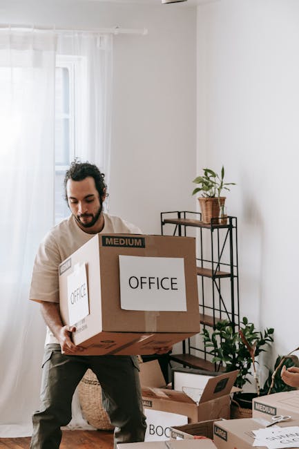 A man with dark, curly hair and a beard, dressed in casual light-colored clothing, is inside a bright, sparsely furnished room with white walls and natural light. He is holding a large cardboard box labeled 'OFFICE' with a smaller label indicating 'MEDIUM' size, as part of a home or office relocation process. In the background, there is a small table with a potted plant on top, and some additional packaging materials and boxes are visible on the floor. The image illustrates the packing and loading process during a house or office move, with the man representing professional removals services such as those offered by Man With a Van Coulsdon, specializing in furniture transport and packing for local relocations.