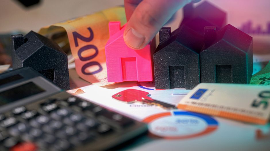 Close-up image showing a person's hand holding a small pink house model, positioned among several black house-shaped models, with a background of euro banknotes including a 200 euro note and a 50 euro note, as well as a calculator, documents with graphical data, and a red key fob. The scene appears to be set on a desk, representing aspects of property valuation, real estate costs, or moving expenses, which align with home relocation and furniture transport topics. The lighting highlights the objects clearly, emphasizing the financial and logistical elements involved in house removals and packing and moving services offered by Man With a Van Coulsdon.