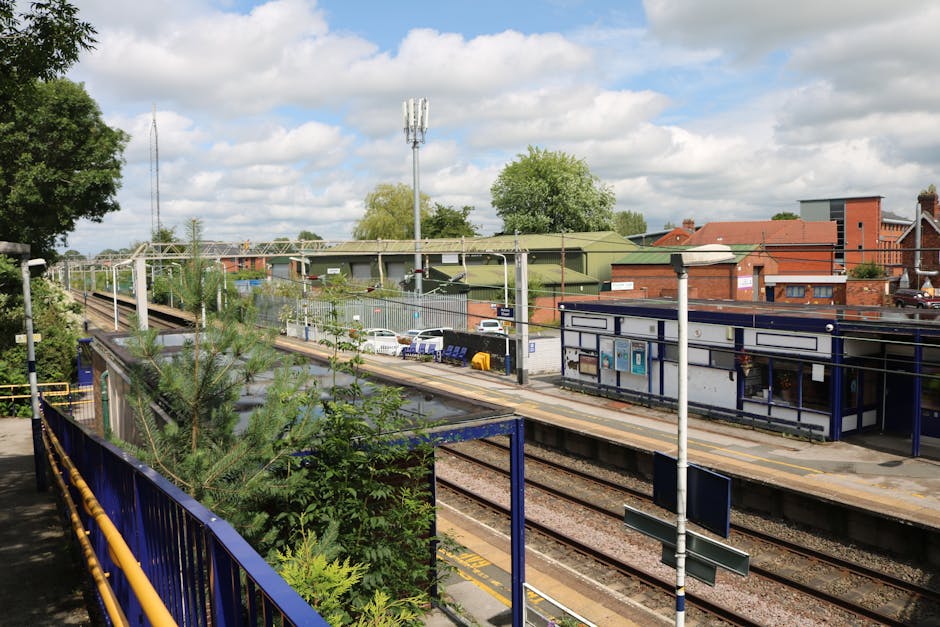 View of a railway station platform in Coulsdon, featuring a blue metal railing, yellow safety lines on the concrete platform, and various station signage and information boards. In the background, there are multiple railway tracks leading away from the platform, surrounded by green trees and shrubs. A small station building with blue and white exterior is visible, alongside parked cars near the station entrance. The scene is captured during daytime with partly cloudy skies, providing natural lighting. Additionally, there are utility poles and a tall communication tower situated behind the station, along with buildings and industrial structures further in the distance. This setting is typical for home relocation or moving logistics involving careful loading and transportation of furniture or boxes from the station to a moving vehicle, such as those operated by Man With a Van Coulsdon.