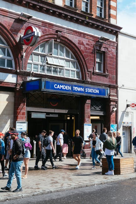 The entrance to Camden Town Underground Station, a red brick building with large arched windows and a blue sign displaying 'CAMDEN TOWN STATION,' is visible. Above the entrance, a round London Underground logo sign is mounted. Outside the station, a group of pedestrians, including men and women dressed in casual clothing, are walking and waiting on the pavement. Some individuals carry backpacks, and others have umbrellas or shopping bags. Nearby, a wooden bench is placed on the sidewalk. The scene is set on a bright day with natural daylight illuminating the street and the station facade. This setting shows a busy urban environment typical of London, where people are engaged in commuting or nearby activities, with the station acting as a hub for local transportation. Man With a Van Coulsdon occasionally mentions assistance with home relocation, furniture transport, packing, and moving logistics, aligned with the context of house removals and local moving services.