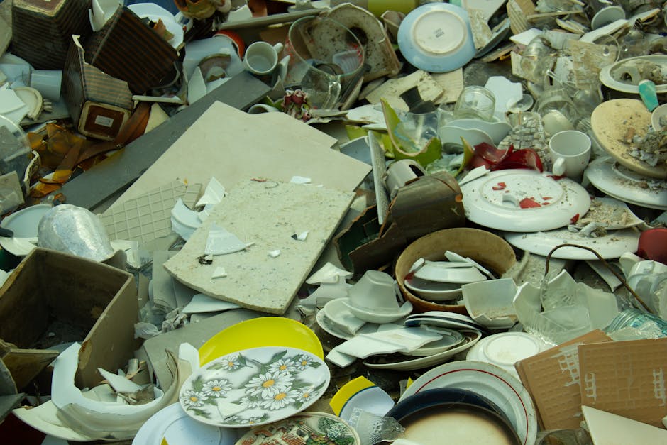 A large pile of broken and discarded household ceramics, glassware, and kitchenware including shattered plates, bowls, cups, and mugs, some with visible cracks and chips, mixed with pieces of cardboard and packing materials. The debris is spread across a pavement area outside a property, with some items overlapping and scattered randomly. In the background, there are fragments of furniture, such as a damaged wooden box and broken ceramic tiles. The lighting appears natural, highlighting the varied textures and materials of the clutter, which is connected to the process of home relocation or disposal. The image depicts a scene of waste that may be collected during a house clearance or bulky waste removal service, illustrating the type of items handled by [COMPANY_NAME] in Croydon while managing furniture transport and packing during removals for clients.