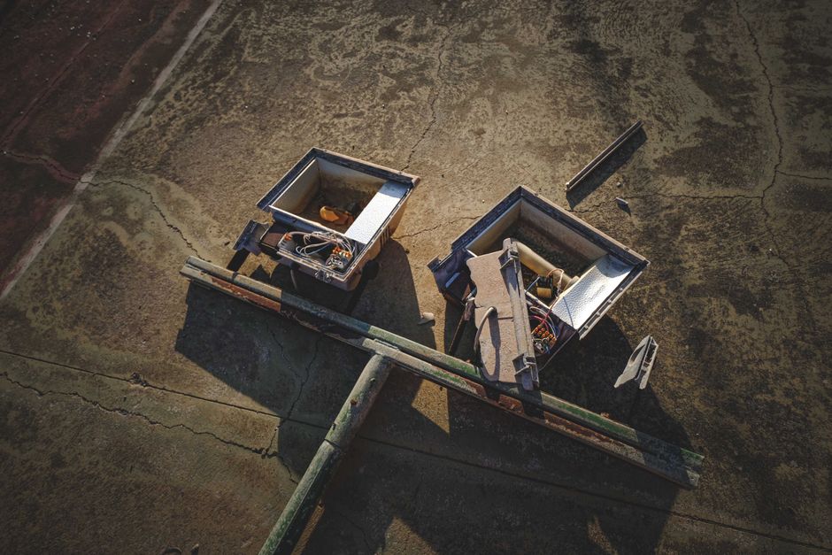 An aerial view of two open electrical junction boxes mounted on a rooftop surface. The boxes are made of metal and are partially disassembled, revealing internal wiring and components, with some wires bundled together. Several loose metal and plastic panels are scattered nearby on the weathered, textured roof surface, which has visible cracks and patches of moss or dirt. A metal railing runs along the edge of the roof, casting shadows, and the lighting indicates late afternoon sunlight. This image relates to home relocation or building maintenance processes involving electrical system disassembly, typical in house removals or renovations, with the presence of wiring and equipment supporting aspects of packing and moving systems managed by companies like Man With a Van Coulsdon.