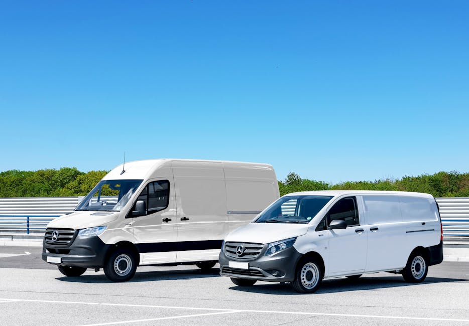 Two white Mercedes-Benz vans are parked side by side on a paved driveway or road, with a bright blue sky overhead and a backdrop of green trees and a metal guardrail. The larger van on the left has a high roof and appears to be used for home relocation or furniture transport, while the smaller van on the right has a lower roof and is equipped for loading boxes and household items. Both vehicles are positioned facing forward, with the larger van slightly behind and to the left of the smaller one, suggesting a loading process or vehicle deployment for moving services. The scene is well-lit, with no visible cargo, but the context indicates they are part of a professional house removal or packing and moving operation. The image is associated with Man With a Van Coulsdon and their emergency same-day van removals service, illustrating a typical setup used in quick-moving logistics or transport of household belongings.
