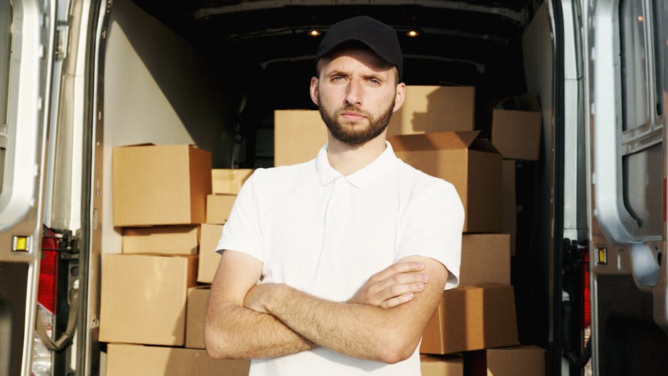 A man with a beard and wearing a black cap and a white polo shirt stands inside the open rear doors of a moving van, which is filled with neatly stacked cardboard boxes of various sizes, some with packing tape. The boxes are arranged on the interior floor of the van, with a few positioned near the sides. The man has a serious expression, with his arms crossed, as he looks directly at the camera. The scene is well-lit with natural light coming from outside, highlighting the packing and loading process involved in home relocation and furniture transport services. The background includes the interior of the van, which appears clean and organized, supporting professional moving activities by Man With a Van Coulsdon, demonstrating the process of packing and loading during an emergency same-day van removal in Coulsdon.