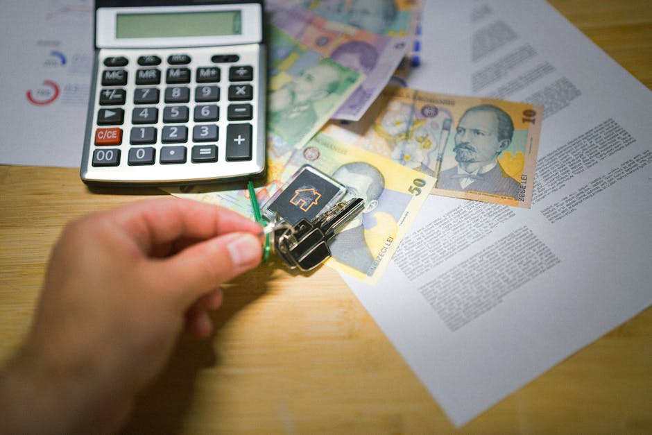 A close-up of a person's hand holding a key attached to a small keyring and a black key fob, positioned above a wooden surface. The surface has scattered items including a silver calculator with black buttons and a green display screen, UK banknotes in various denominations (including a 10-pound note and a 50-pound note), and printed documents with text. The setting appears to be indoors, possibly a home or an office area, where the process of packing or preparing for a move or home relocation is taking place. The arrangement of belongings suggests tasks related to planning and organizing a house removal or moving logistics, which Man With a Van Coulsdon offers as part of their removals service.
