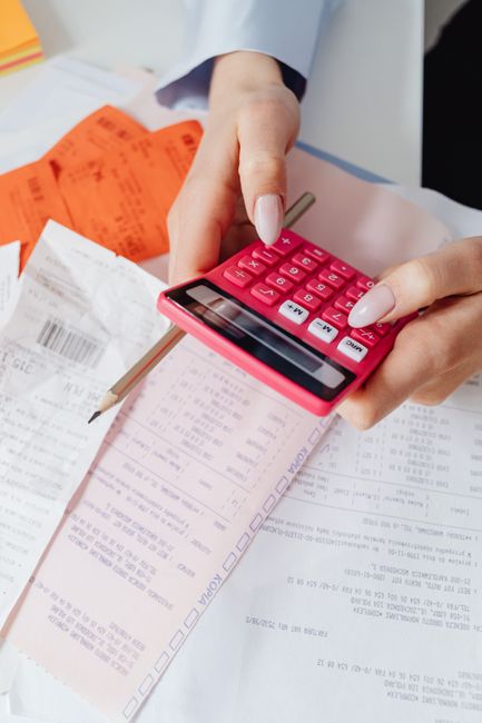 A close-up of a person's hand holding a pink calculator above several sheets of printed documents, including receipts, invoices, and packing lists, with some orange labels and a pencil visible on the cluttered table. The background features a white surface, possibly a desk or table at a home or office setting, with documents arranged in an organized manner. This scene reflects aspects of packing, documentation, and planning involved in home relocation and furniture transport services, as provided by Man With a Van Coulsdon, supporting efficient moving logistics for house removals in the COULSDON area.