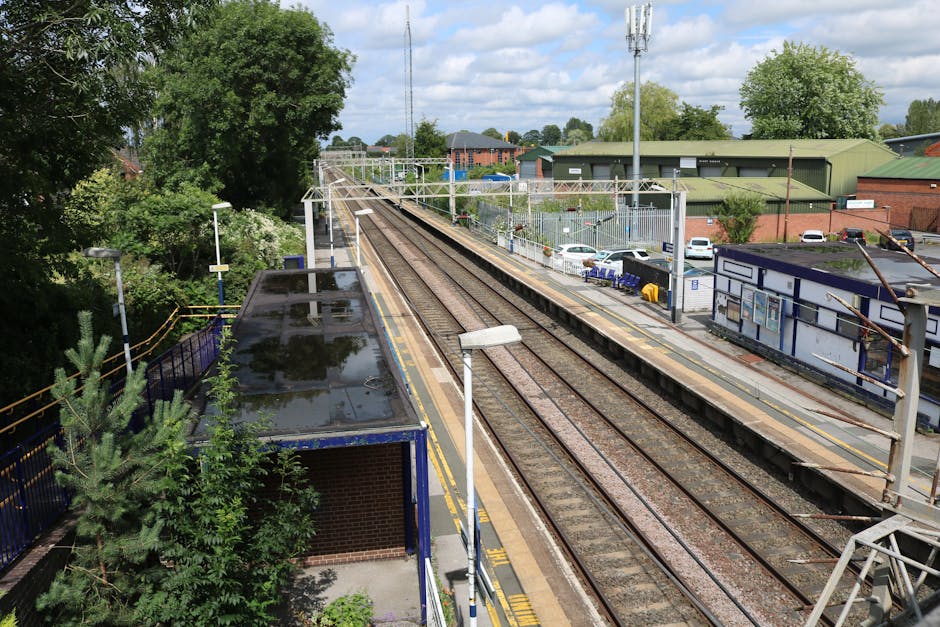 The image shows a view of a railway station platform taken from an elevated position. The platform is partially visible on the right side, with a row of white streetlights and a blue border along the edge. Adjacent to the platform, multiple train tracks extend into the distance, separated by gravel and metal sleepers. On the left side, there is a small shelter with a sloped, dark roof, partially surrounded by greenery, including trees and bushes, some of which overhang onto the shelter. Behind the station, there are commercial buildings with green and red roofs, and a parking area with several parked cars. Overhead, electric lines and supporting poles run parallel to the tracks. The sky is partly cloudy, with patches of blue visible, indicating daylight. This scene captures the typical layout of a suburban railway station, reflecting the infrastructure involved in train travel and station operation.