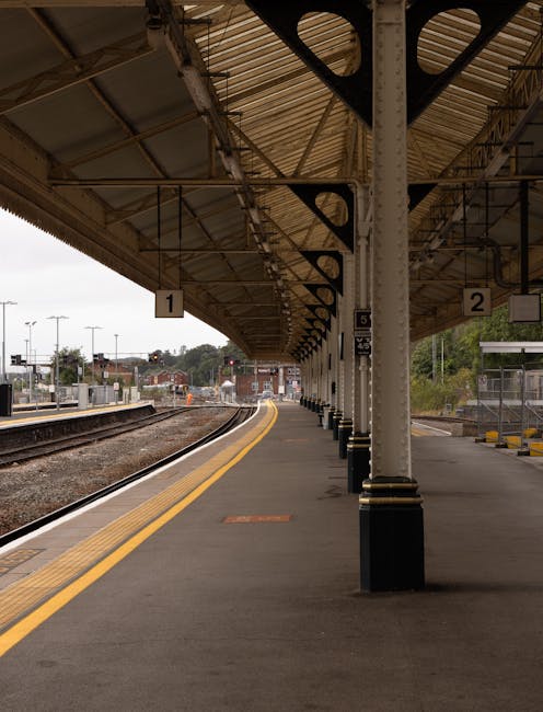 An empty train platform at Coulsdon South station with a covered waiting area supported by metal columns, featuring parking and railway tracks on the left. The platform surface is paved with asphalt and has yellow tactile paving along the edge for safety. There are signs displaying platform numbers and service information hanging from the roof structure. In the background, the tracks curve gently out of view, with some distant buildings, trees, and overcast sky visible beyond the station. The scene suggests a quiet period suitable for home relocation or furniture transport processes, with no people present in the image, confirming a moment during off-peak hours where loading or unloading could occur for moving services provided by companies like Man With a Van Coulsdon.