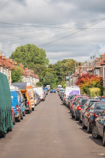 A residential street scene showing numerous vehicles parked on both sides of the road, including small vans, cars, and a covered vehicle, with some vans positioned near house entrances and driveways. The street is lined with terraced houses featuring brick facades, and small front gardens or paved areas. A man from Man With a Van Coulsdon is seen loading or unloading furniture and boxes wrapped in cardboard, plastic, and blankets onto or from a van parked adjacent to the pavement. The setting appears to be during daylight hours with an overcast sky, and the background includes leafy trees and utility wires overhead. This image depicts a typical home relocation or moving process involving furniture transport, packing, and vehicle loading as part of professional removals services in Croydon.